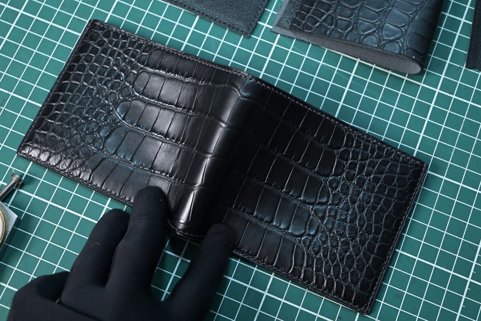 Gloved hands holding open a black crocodile leather wallet on a green cutting mat